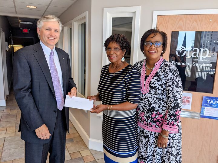Three people standing together looking at the camera. One man hands a check to one woman
